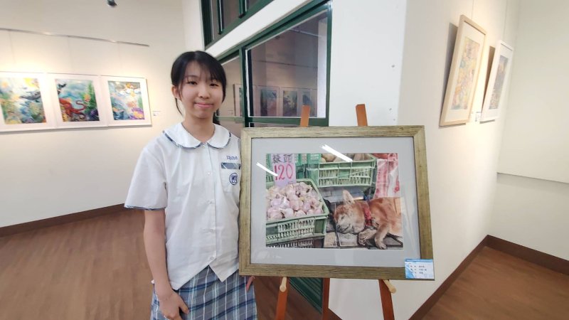 學生謝可柔則以西畫作品「市場一隅」描繪清晨早市的生活場景。攤位桌腳下，一隻柴犬安靜熟睡，形成熱鬧與寧靜之間的鮮明對比，展現學生對生活細節的細膩觀察。（漾新聞記者陳雯萍攝）