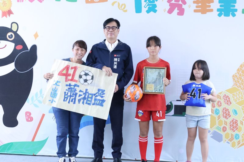 文山國小畢業生蕭湘盈（右二）穿著學校足球隊服與市長合影，她曾與隊友勇奪全國女子足球賽季軍，母親也驕傲地手持印有女兒姓名的應援海報，共同見證這份榮耀。